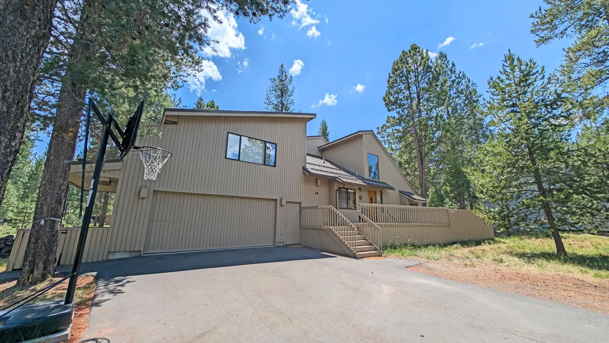 The image displays the home's front entrance, framed by mature trees, and features a basketball hoop, hinting at active outdoor fun. The welcoming pathway and spacious area in front suggest easy access to Sunriver's extensive bike paths and outdoor adventures.