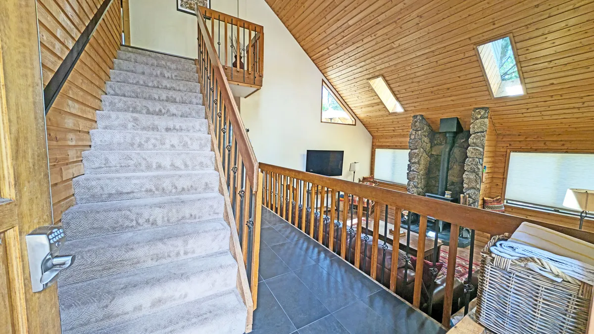 A warm, open living area featuring a spacious staircase with wooden railings, a striking stone fireplace, and expansive windows that frame the natural surroundings. The room is arranged to encourage group gatherings, with ample seating and a cozy atmosphere ideal for relaxing, socializing, or enjoying views outside.