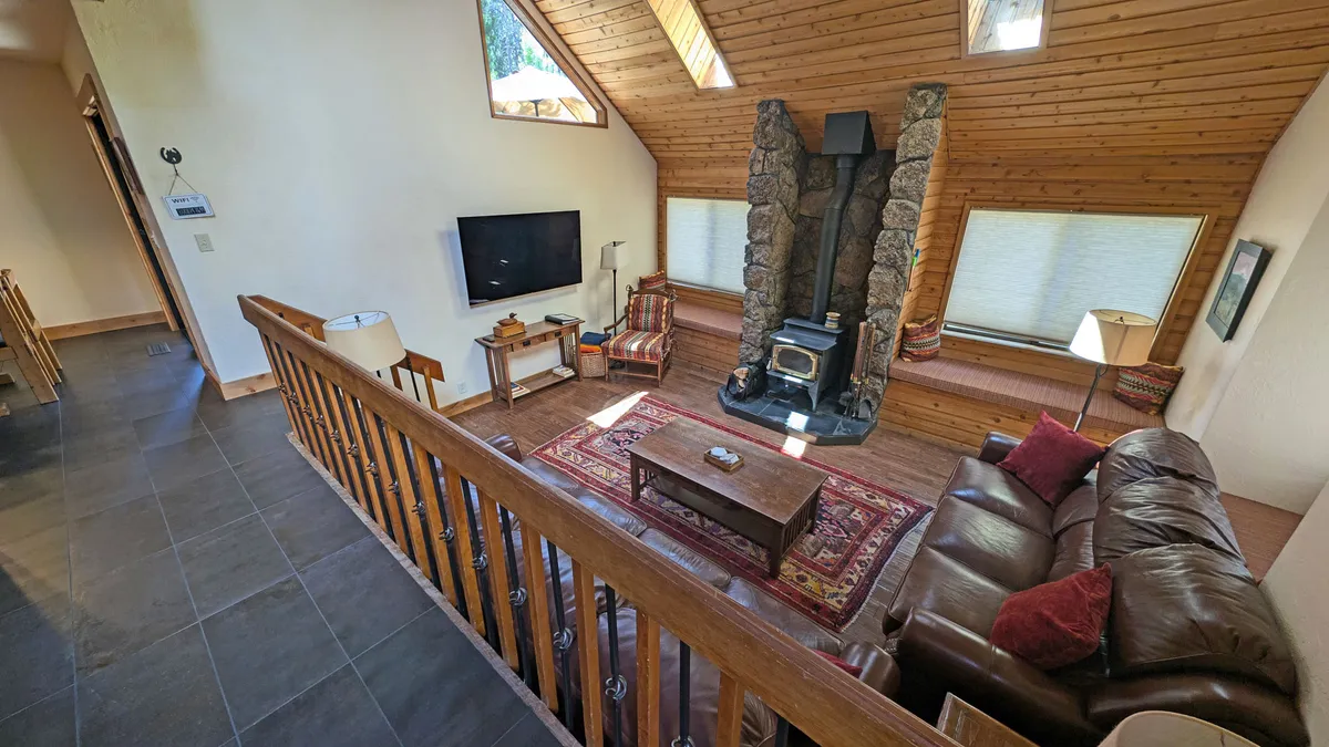 A welcoming living room anchored by a stone fireplace, with a plush leather sofa, exposed wooden beams in the ceiling, and a patterned rug. The space exudes warmth and is flooded with natural light from generous windows, creating an ideal setting for cozy gatherings, movie nights, or relaxed conversations.