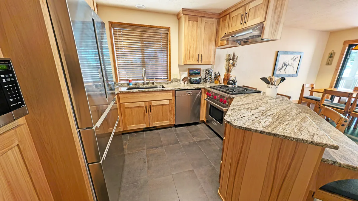 A warm kitchen scene with granite countertops, stainless steel appliances, and wood cabinetry. The window with blinds suggests a connection to the outdoors, and the ambiance supports group cooking and entertaining.