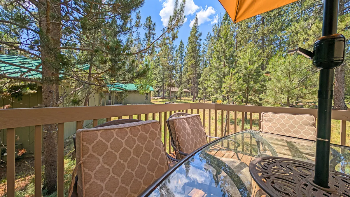 A peaceful outdoor dining area with a glass table and decorative chairs sits under a large umbrella. The seating is set against a backdrop of mature trees, evoking the feeling of being close to nature and offering a perfect transition zone between indoors and outdoors.