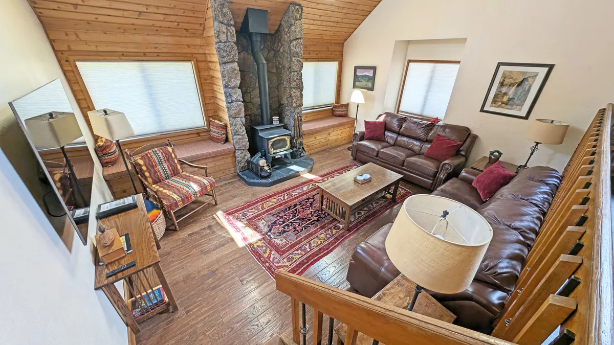 An inviting interior scene centered on a stone fireplace, surrounded by leather sofas and rustic wooden furniture. The colorful area rug and warm lighting create a welcoming space perfect for shared meals or quiet conversation after a day of adventure.